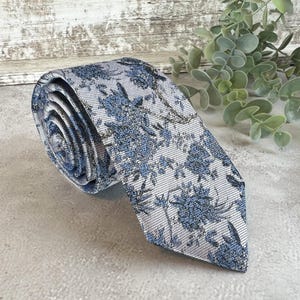 May include: Rolled-up necktie with a floral pattern in shades of blue and gray. The tie is made of a textured fabric and features a repeating design of stylized flowers and leaves. The tie is on a light gray surface.