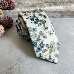 Könnte beinhalten: Eine weiße Krawatte mit einem floralen Muster aus blauen und grünen Blumen und Blättern. Die Krawatte ist gerollt und zeigt das detaillierte Design. Der Hintergrund ist hellgrau.