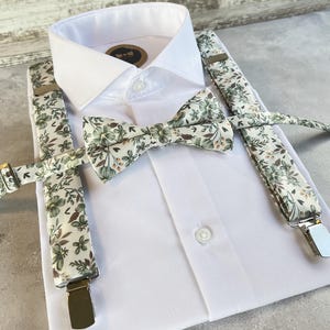 Può includere: Una camicia bianca con papillon e bretelle abbinati. Il papillon e le bretelle presentano uno sfondo color crema con motivi floreali verdi e marroni. Le bretelle hanno clip metalliche argentate.