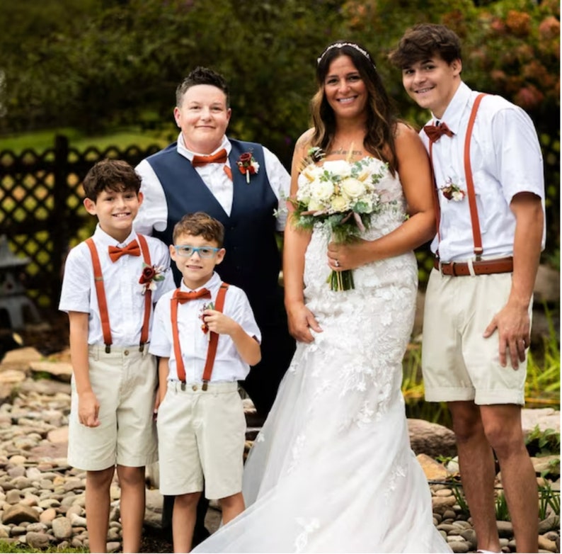 Op de afbeelding: Een bruiloftsfeest met twee bruiden, twee jonge jongens in bijpassende outfits en een bruidegom. De bruiden dragen witte jurken, de jongens dragen witte overhemden met oranje strikjes en bretels, en de bruidegom draagt een wit overhemd met een oranje strik en bretels. De bruiden houden een boeket witte bloemen vast.