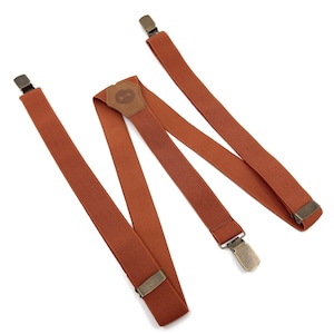 Op de afbeelding: Een paar bruine bretels met messing clips en een leren patch met een striklogo.