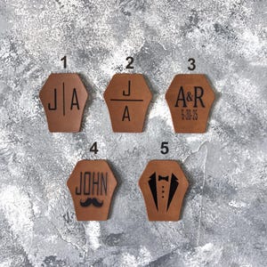 Op de afbeelding: Vijf bruine leren patches met verschillende ontwerpen. Patch 1 heeft de initialen "J|A". Patch 2 heeft de initialen "J/A". Patch 3 heeft de initialen "A&R" en de datum "6-30-25". Patch 4 heeft de naam "JOHN" en een snor. Patch 5 heeft een smoking met een strikje.