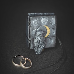 May include: A black leather-bound journal with a raven and crescent moon design. The journal is closed and has a black cord tied around it. Two gold wedding bands are in front of the journal.