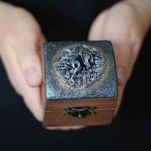Op de afbeelding: Een klein houten doosje met een scharnierend deksel en een metalen sluiting. Het doosje is versierd met een zilveren Keltisch knoopontwerp en een zilveren wolfskop.