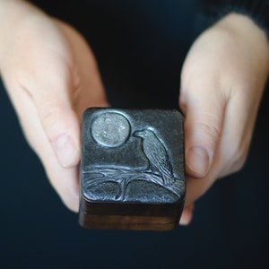 Peut inclure: Une petite boîte en bois avec un dessus en métal argenté. Le dessus présente un motif gravé d'un corbeau perché sur une branche avec une pleine lune en arrière-plan.