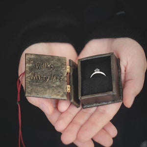 Op de afbeelding: Een klein, bruin, lederen doosje met de woorden "Will You Marry Me?" gegraveerd op de voorkant. Het doosje is open en laat een zilveren ring zien met een enkele diamant in het midden.