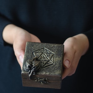 May include: A wooden box with a gold-toned lid featuring a 20-sided die design and a sculpted leaf detail. The box has a brass latch.