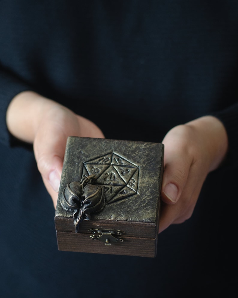 May include: A wooden box with a gold-toned lid featuring a 20-sided die design and a sculpted leaf detail. The box has a brass latch.