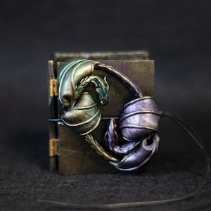 May include: A small black leather-bound journal with a sculpted dragon design on the front cover. The dragon is made of a green and purple iridescent material and has a long tail that wraps around the journal. The journal is attached to a black cord.