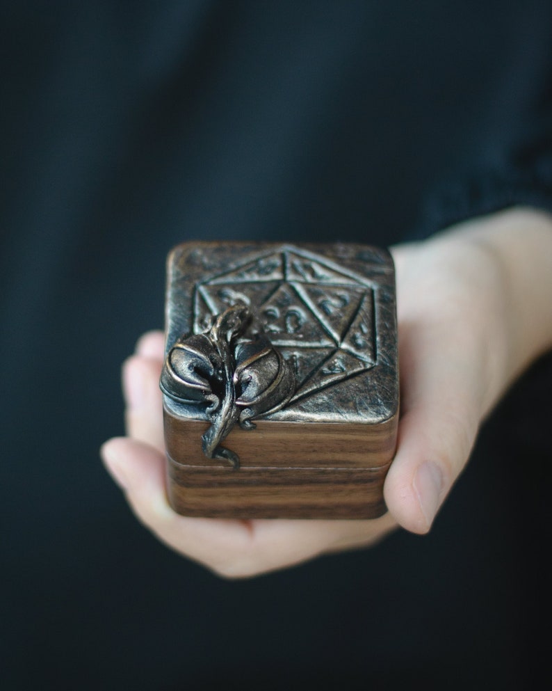 May include: A wooden box with a metal top featuring a stylised 20-sided die design. The box has a metal leaf design on the side.