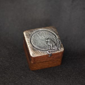 May include: A small, wooden box with a silver-toned metal lid featuring a detailed relief of a dragon in front of a full moon. The box has a rectangular shape with a dark brown finish and a silver-toned metal border around the lid.