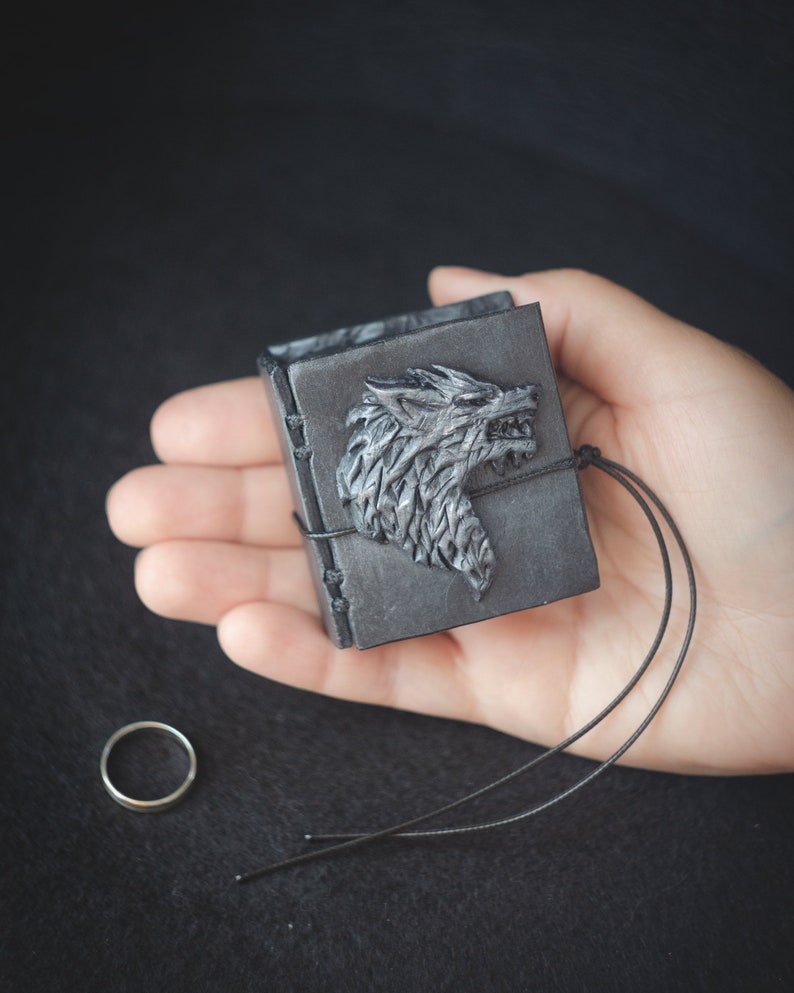 May include: A small black leather-bound book with a silver wolf head design on the cover. The book is closed and has a black cord attached to it.