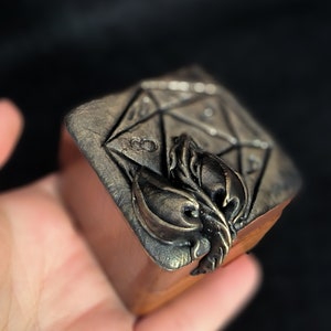 May include: A small wooden box with a metal design of a 20-sided die and a stylised leaf on the top.