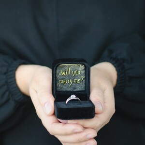 Pode incluir: Uma caixa de anel de veludo preto com as palavras "Will you marry me?" em letras douradas. Um anel de prata com um &uacute;nico diamante est&aacute; dentro da caixa.
