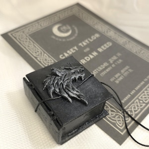 May include: A black book-shaped box with a silver wolf head design on the front. The box is tied with a black cord and is sitting on a gray card with text that reads "Casey Taylor and Jordan Reed".