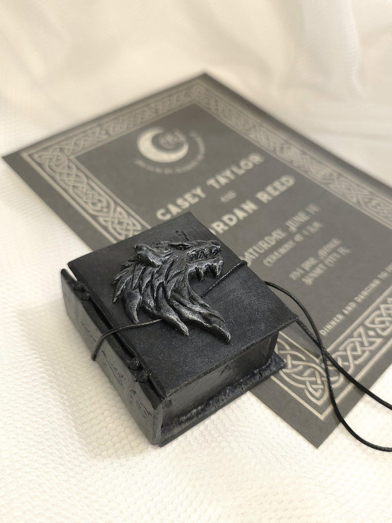 May include: A black book-shaped box with a silver wolf head design on the front. The box is tied with a black cord and is sitting on a gray card with text that reads "Casey Taylor and Jordan Reed".