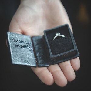 May include: A black ring box with the words "You are my everything" on the front. The box is open and a silver ring with a single diamond is inside.