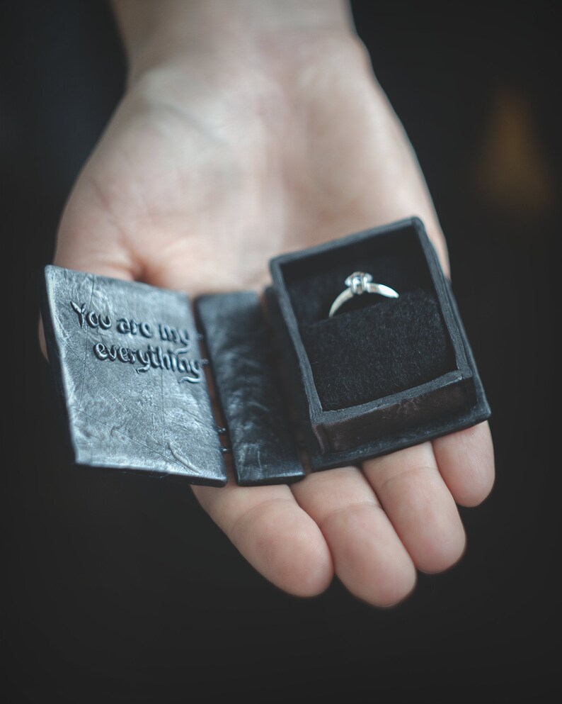 May include: A black ring box with the words "You are my everything" on the front. The box is open and a silver ring with a single diamond is inside.