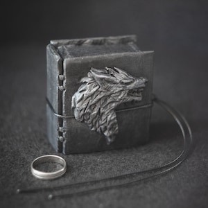 May include: A black leather ring box with a wolf head design on the front. The box is closed and has a black leather strap.