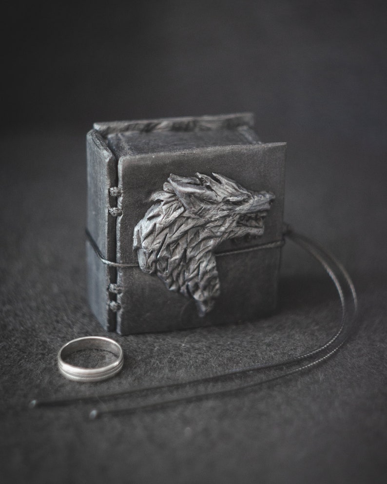 May include: A black leather ring box with a wolf head design on the front. The box is closed and has a black leather strap.