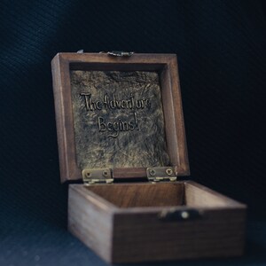 May include: A wooden box with a hinged lid. The inside of the lid is lined with a textured, gold-coloured material. The lid is open and the inscription "The Adventure Begins!" is visible. The box is made of dark wood and has a brass latch.