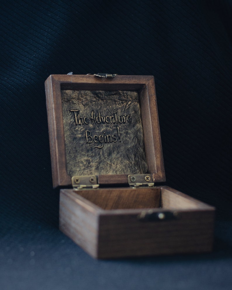 May include: A wooden box with a hinged lid. The inside of the lid is lined with a textured, gold-coloured material. The lid is open and the inscription "The Adventure Begins!" is visible. The box is made of dark wood and has a brass latch.