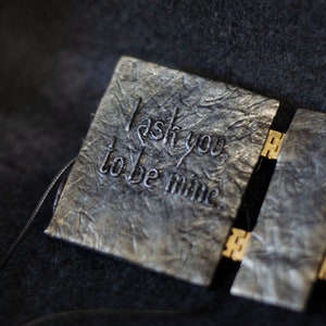 K&ouml;nnte beinhalten: Ein kleines, schwarzes, ledergebundenes Buch mit einem goldenen Verschluss. Der Einband hat die Worte "Ich bitte dich, mein zu sein" in Silber gepr&auml;gt.