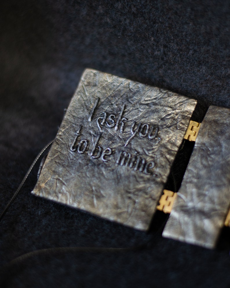 K&ouml;nnte beinhalten: Ein kleines, schwarzes, ledergebundenes Buch mit einem goldenen Verschluss. Der Einband hat die Worte "Ich bitte dich, mein zu sein" in Silber gepr&auml;gt.