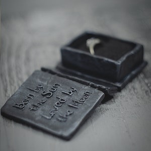 Op de afbeelding: Een zwarte ringdoos met de inscriptie "Born by the Sun Loved by the Moon". De doos is open en er ligt een zilveren ring in.