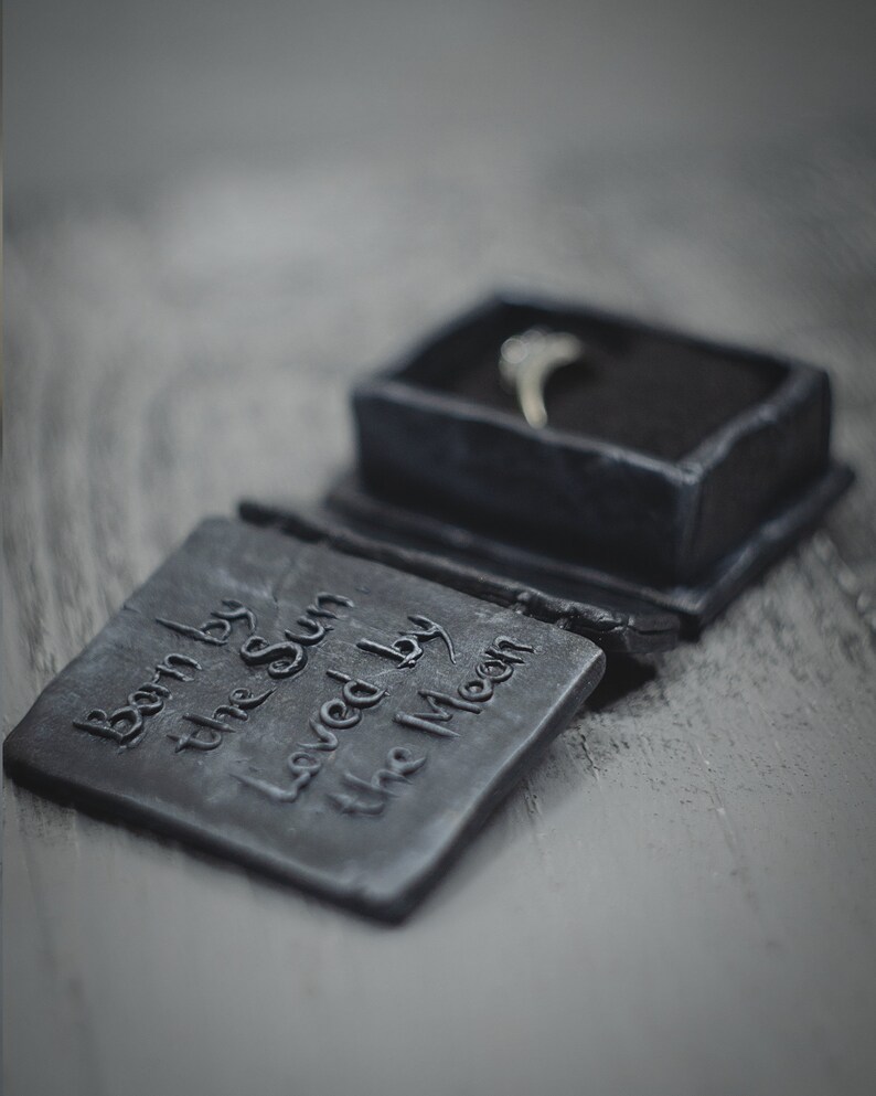 Op de afbeelding: Een zwarte ringdoos met de inscriptie "Born by the Sun Loved by the Moon". De doos is open en er ligt een zilveren ring in.
