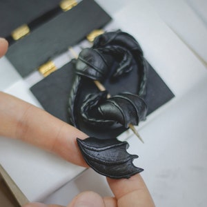 K&ouml;nnte beinhalten: Ein schwarzer Drachenfl&uuml;gel-Anh&auml;nger aus Polymer Clay. Der Fl&uuml;gel ist mit strukturierten Schuppen verziert und an einer kleinen Nadel befestigt.