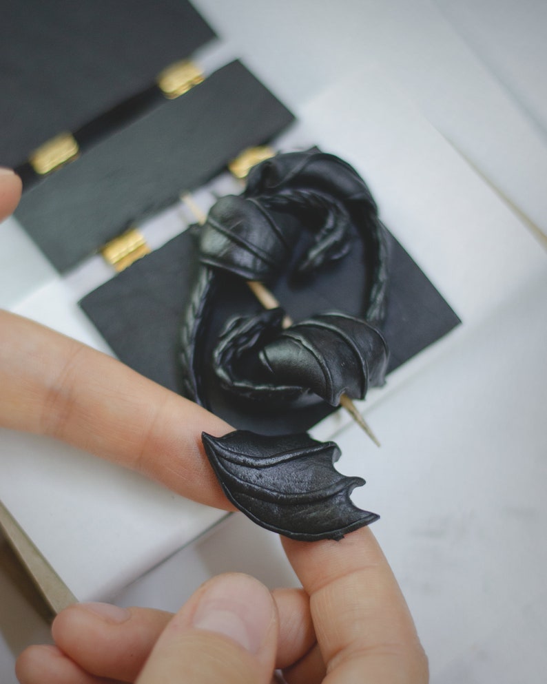 K&ouml;nnte beinhalten: Ein schwarzer Drachenfl&uuml;gel-Anh&auml;nger aus Polymer Clay. Der Fl&uuml;gel ist mit strukturierten Schuppen verziert und an einer kleinen Nadel befestigt.