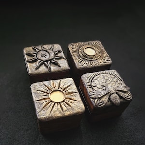 May include: Four small square boxes with different designs. Each box has a gold-colored finish and a raised design. The designs include a sun with a crescent moon, a sun with a circle in the center, a honeycomb with a bee, and a sun with a circle in the center.