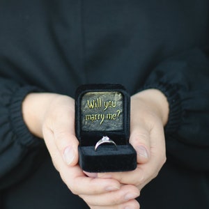 May include: A black velvet ring box with the words "Will you marry me?" in gold lettering. A silver ring with a single diamond is inside the box.