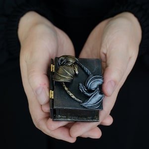 K&ouml;nnte beinhalten: Eine kleine, schwarze Holzkiste mit einem Scharnierdeckel. Die Kiste ist mit zwei Drachen verziert, einem goldenen und einem silbernen, die mit einem Seil ineinander verschlungen sind.