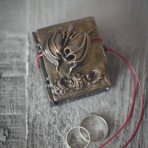 Op de afbeelding: Een klein, versierd zwart leren doosje met een goudkleurig metalen drakenontwerp op de deksel. Twee zilveren trouwringen liggen naast het doosje op een grijs houten oppervlak.