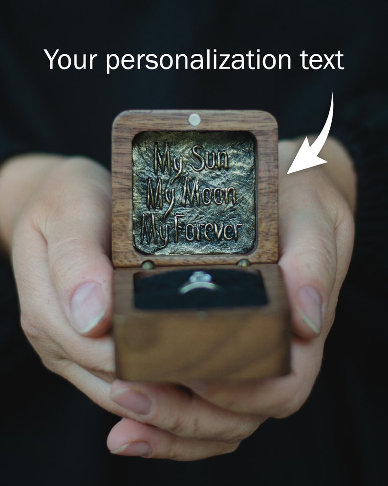 May include: A wooden ring box with a black velvet interior. The lid is open and has a gold-coloured inscription that reads "My Sun My Moon My Forever".