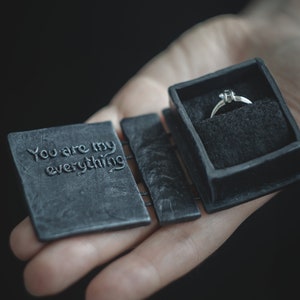 May include: A black ring box with the words "You are my everything" engraved on the lid. The box is open and a silver ring with a single diamond is inside.