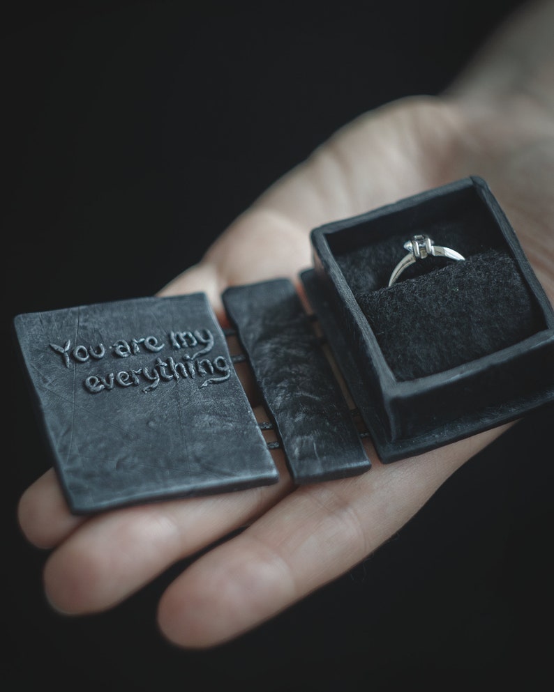 May include: A black ring box with the words "You are my everything" engraved on the lid. The box is open and a silver ring with a single diamond is inside.