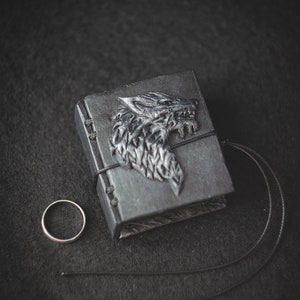 May include: A black leather-bound journal with a silver wolf head design on the cover. A silver ring is next to the journal.