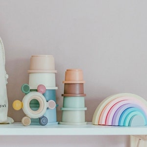 May include: A selection of pastel-coloured baby toys, including stacking cups, a rainbow arch, and a teether. The toys are arranged on a white shelf against a soft pink background. A canvas bag is visible on the left.