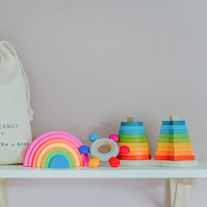 May include: A rainbow-colored wooden arch, a colorful wooden stacking toy, and a blue and green wooden stacking toy on a white shelf. The stacking toys are in the shape of stars. The shelf is against a light pink wall.