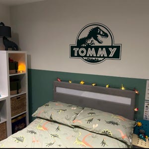 Op de afbeelding: Een groene en witte slaapkamer met dinosaurusthema, met een bed, een witte boekenkast en een dinosaurusmuursticker met de tekst "TOMMY".
