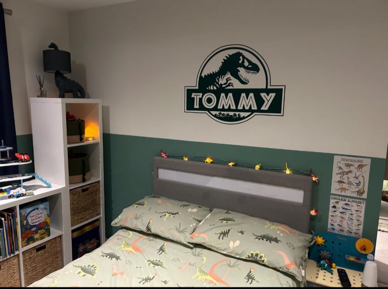 Op de afbeelding: Een groene en witte slaapkamer met dinosaurusthema, met een bed, een witte boekenkast en een dinosaurusmuursticker met de tekst "TOMMY".