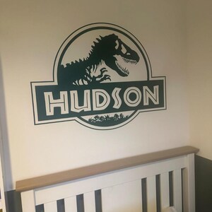 Op de afbeelding: Groene dinosaurus silhouet met de tekst "HUDSON" in een witte omtrek, binnen een groene cirkel met een groene achtergrond. De afbeelding is een muursticker.