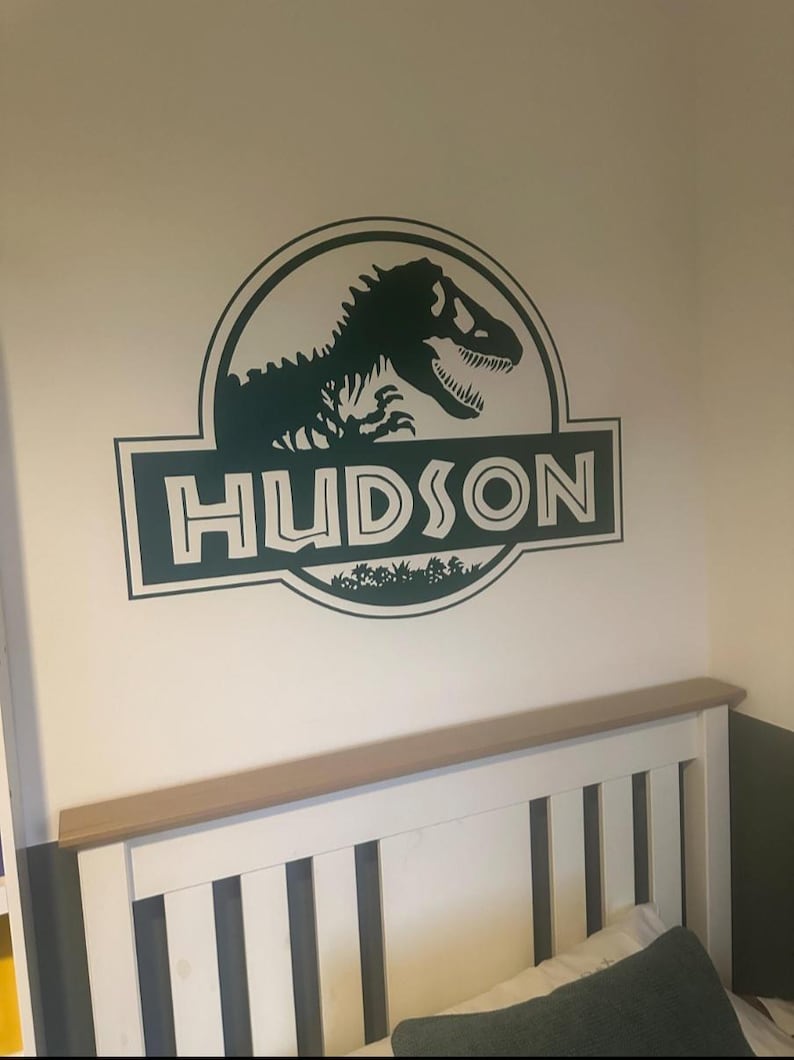 Op de afbeelding: Groene dinosaurus silhouet met de tekst "HUDSON" in een witte omtrek, binnen een groene cirkel met een groene achtergrond. De afbeelding is een muursticker.