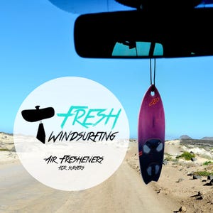 Puede incluir: Un ambientador de aire en forma de tabla de windsurf rosa y negra colgado del espejo retrovisor de un coche. El ambientador tiene el texto "JP" en él. El coche está conduciendo por un camino de tierra con dunas de arena en el fondo. El logotipo "Fresh Windsurfing Air Fresheners for Surfers" es visible en primer plano.