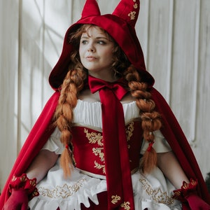 May include: A red hooded cape with gold embroidery and a white lace trim. The hood has cat ears and a red satin bow. The cape is worn over a white dress with a red bodice.