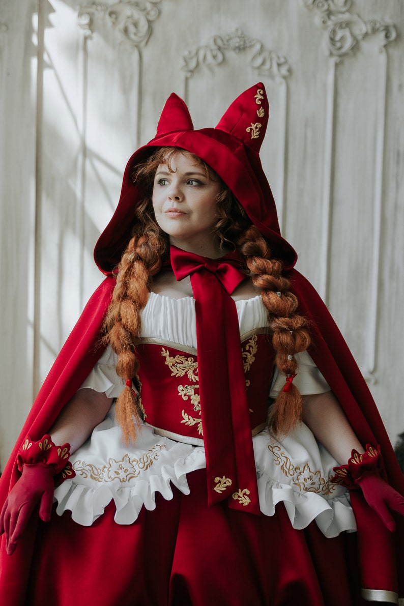 May include: A red hooded cape with gold embroidery and a white lace trim. The hood has cat ears and a red satin bow. The cape is worn over a white dress with a red bodice.