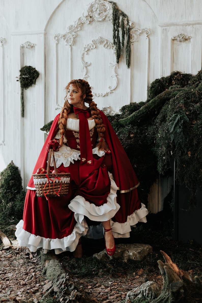 May include: A woman dressed as Little Red Riding Hood, wearing a red hooded cape, a white lace collar, and a red and gold corset. She is holding a basket of red apples and is standing in a forest setting.